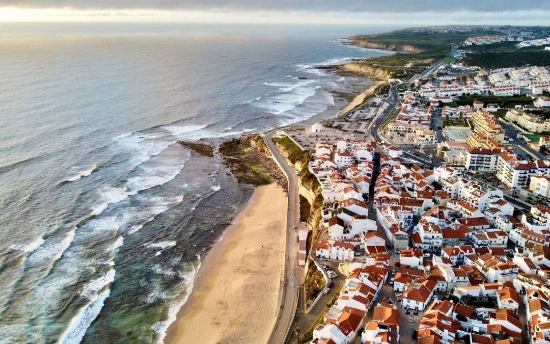Ericeira from the air at sunrise. White houses with terracotta roofs sit on the clifftop above a beach and the Atlantic swell.