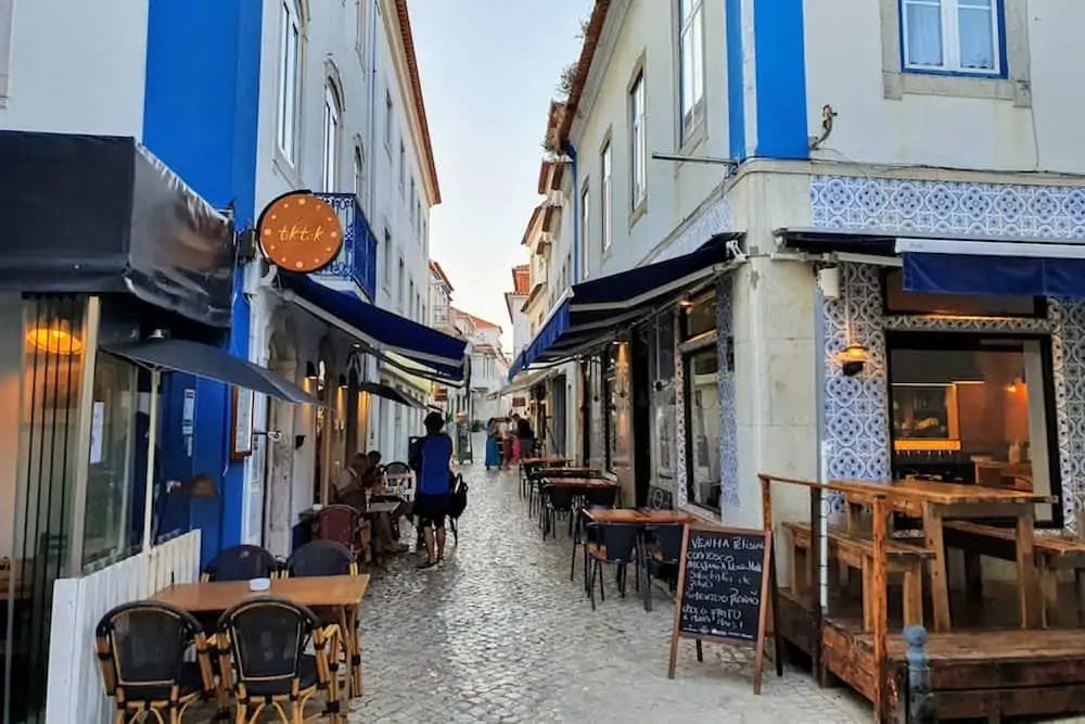 A cobbled street in the old part of Ericeira. People sit at café tables under blue awnings beside tiled house fronts.